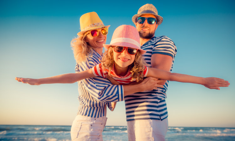 Couple at the beach wearing striped blue shirts holding their daughter in a red striped shirt, with hats and sunglasses