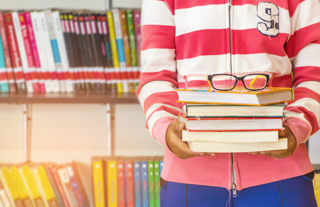 College freshman student holds a stack of textbooks and their glasses while standing in front of a library shelf
